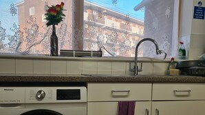 Una cuina amb rentadora, una finestra amb cortina floral i vista d'un edifici a l'exterior.
