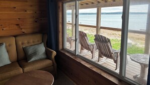 A wooden cabin interior with a sofa, a small table, and a large window overlooking a wooden deck and a lake.