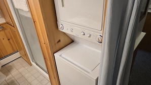 Une machine à laver compacte, de couleur blanche, dotée de plusieurs boutons de commande et d’un petit tiroir de rangement, installée dans un espace étroit aux murs en bois et au sol carrelé.