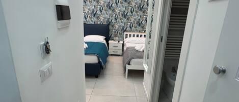 En smal gang der fører til et soveværelse med tapet med et mønster, en seng med himmelblåt sengegavl, et sengebord og en dør med en QR-kode.
