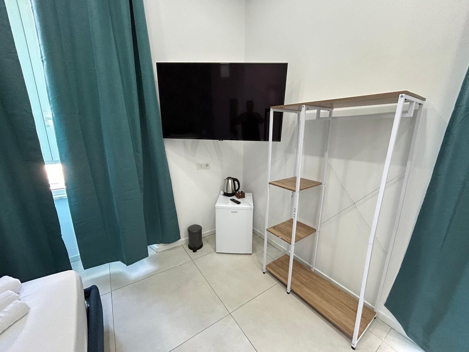 A small, modern room with a bed, a TV mounted on the wall, a coffee maker, and a wooden shelf unit.