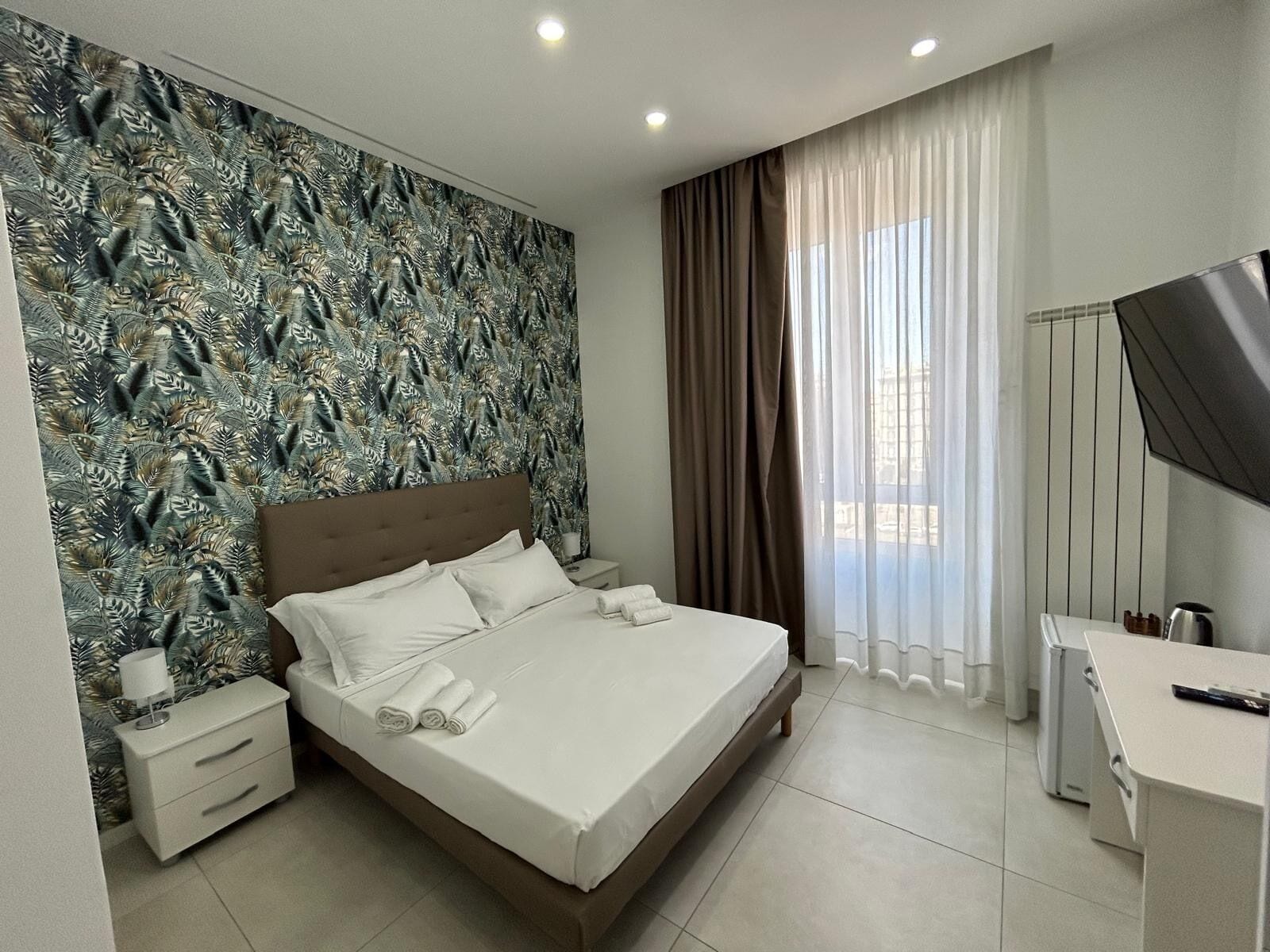 A modern hotel room with a large bed, a desk, a TV, and a window with curtains.
