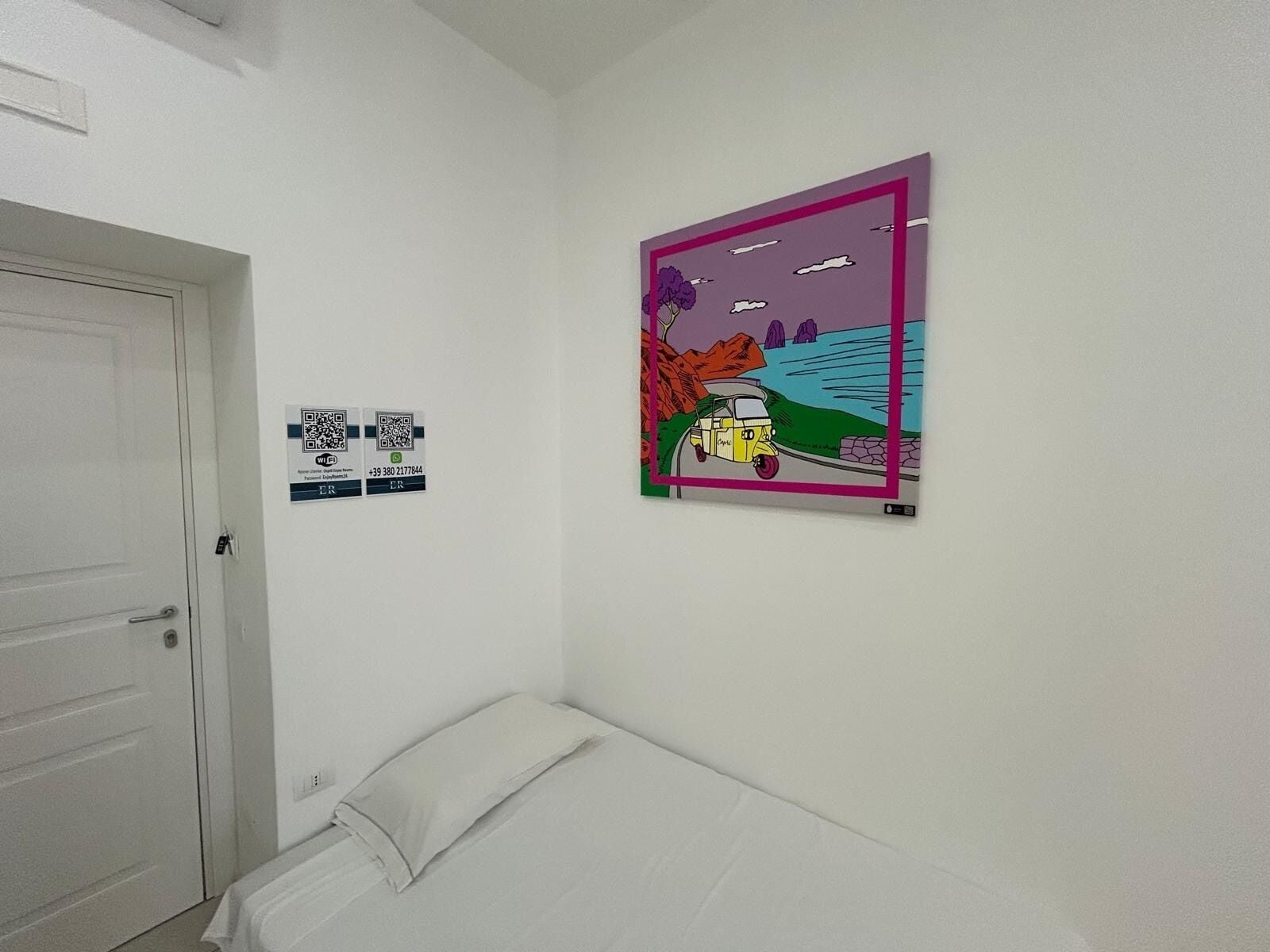 Een kamer met een bed, een deur en een kunstwerk aan de wand dat een landschap met een voertuig toont.