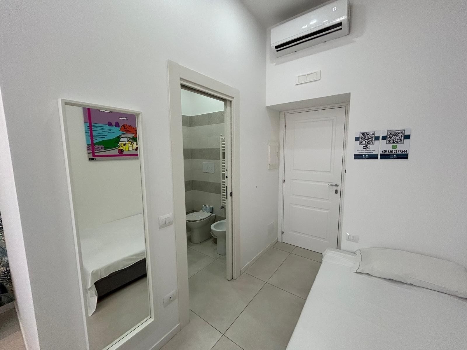 Una habitació d'hotel amb un llit, un bany amb vàter i dutxa, i una porta que dona a una altra habitació.