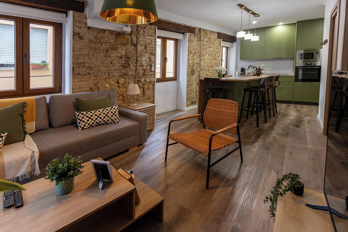 A modern living room with a sofa, armchair, and coffee table, a kitchen with green cabinets, and a stone wall.