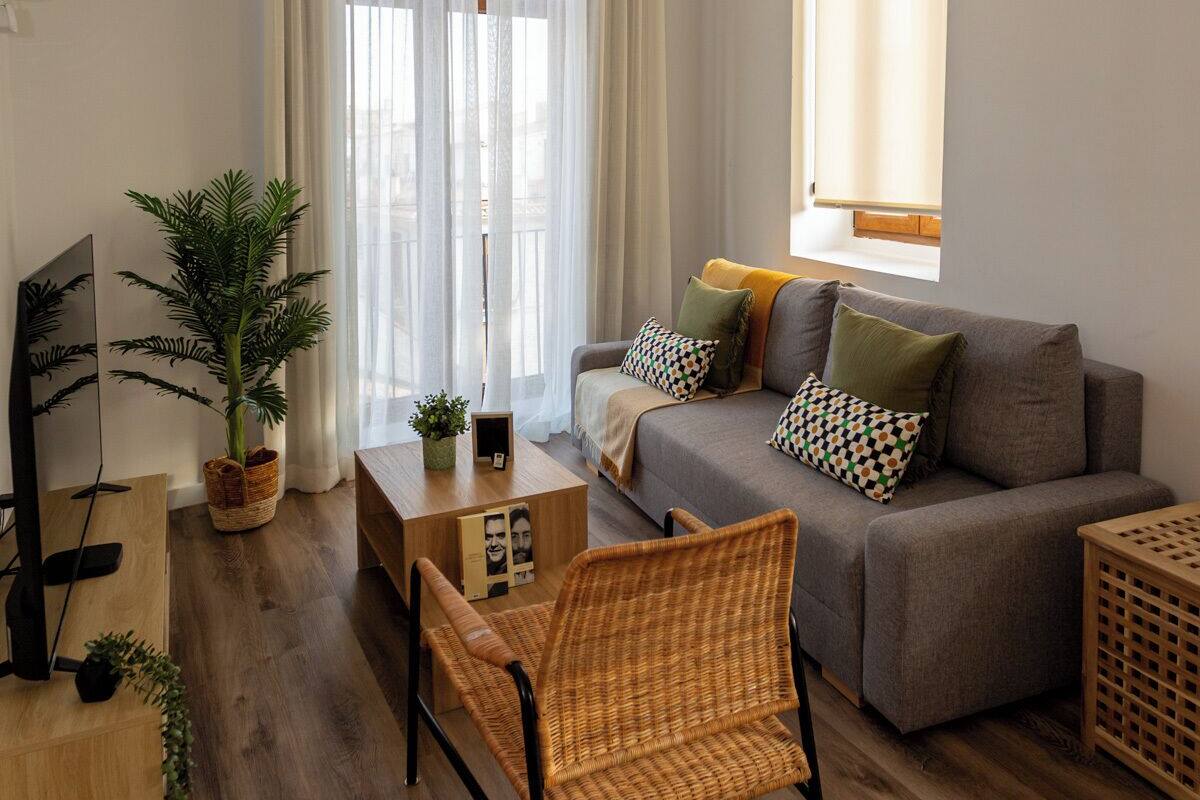 A modern living room with a grey sofa, a wooden coffee table, and a wicker chair. There is a large window with sheer curtains, a hanging green pendant light, and a potted plant.