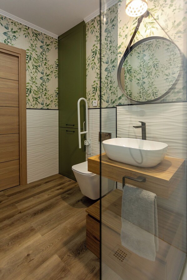 Un baño moderno con piso de madera, inodoro blanco, tocador de madera con lavamanos blanco y cabina de ducha de vidrio.