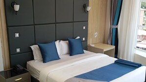 A modern hotel room with a large bed, wooden panel wall, and a view of the outside. - Zuri Place Hotel Limited (Oyugis)