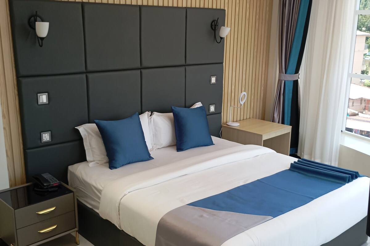 A modern hotel room with a large bed, wooden panel wall, and a view of the outside.