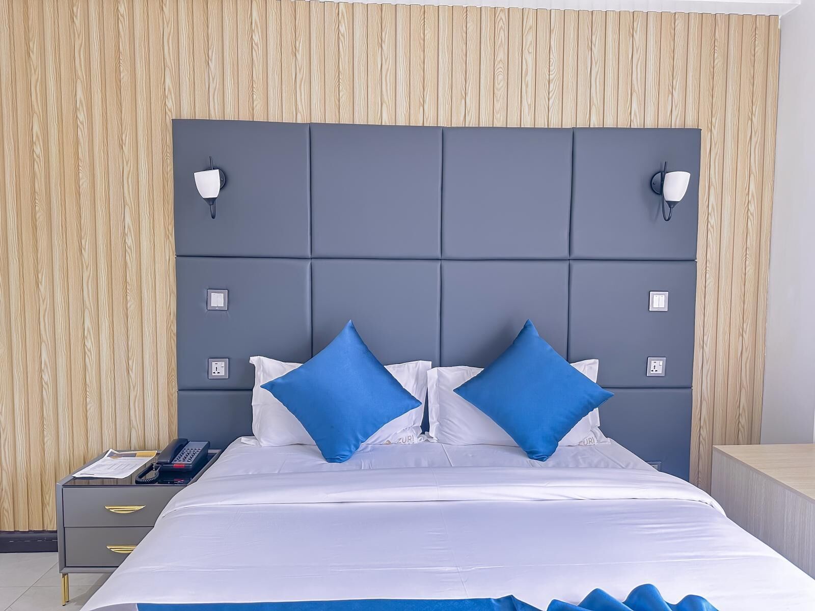 Una habitació d'hotel moderna amb un capçal de llit gris, roba de llit blanca i dos coixins blaus decoratius.