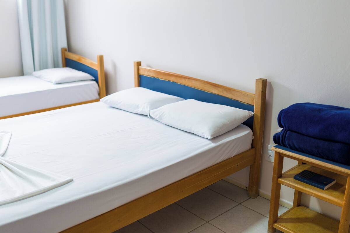 A room with two single beds, each with white bedding and wooden headboards. There is a wooden nightstand with a blue towel and a book on it.