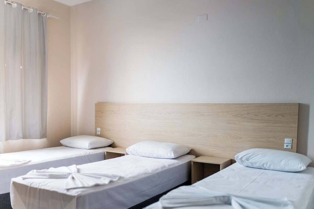 A hotel room with two single beds, a wooden headboard, a small bedside table, and a window with curtains.