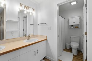 Un baño con tocador de madera, dos lavamanos, un espejo grande con iluminación, un inodoro y un armario con una planta.