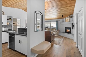 Una cocina moderna con armarios blancos, lavavajillas y un banco empotrado. Se ve un área de estar con chimenea y sofá a través de un umbral abierto.