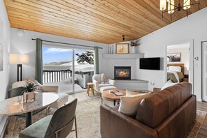 Una sala de estar moderna con chimenea, televisor y balcón con vista a colinas nevadas.