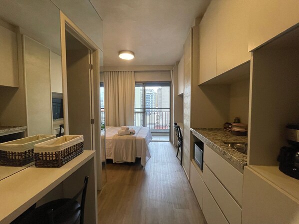 En smal hotelgang med en seng, et badeværelse med håndvask og spejl, samt en balkon med udsigt over byen.