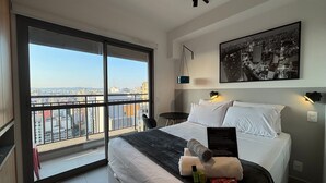 A hotel room with a large bed, a city view through a sliding door, a framed cityscape picture, and a small table with a chair. - Prime Stay - Vila Buarque - São Paulo - BR (São Paulo)