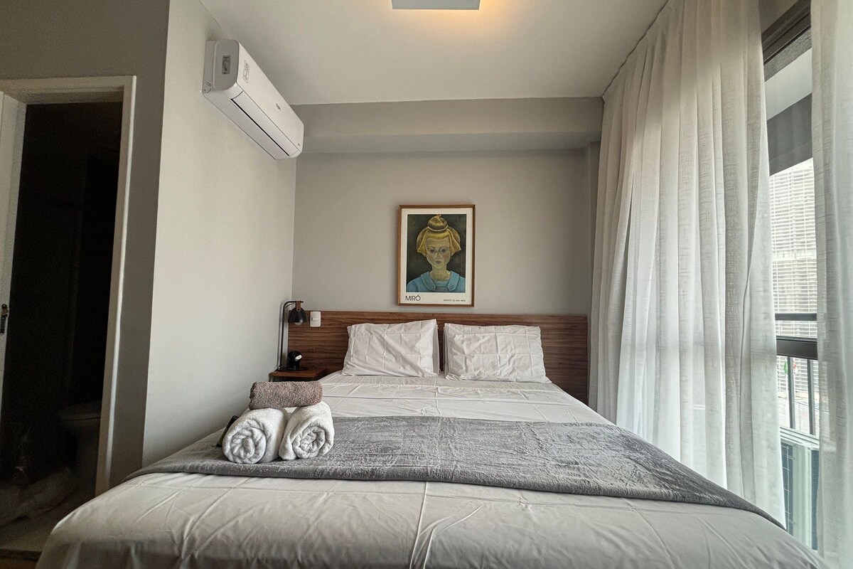 A hotel room with a bed, a painting on the wall, a window with curtains, and an air conditioning unit.