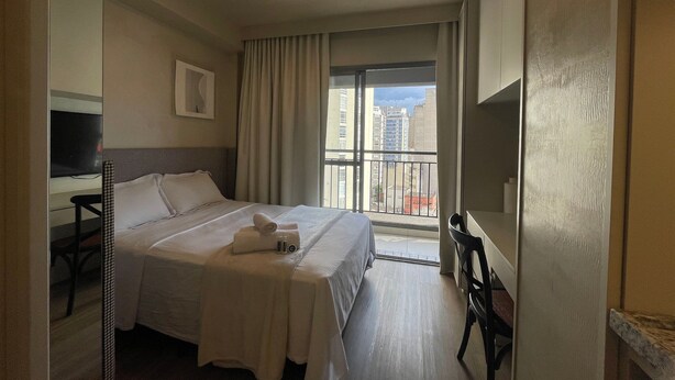Hotelkamer met een bed, een televisie, een stoel, een balkon met uitzicht op gebouwen en een plafondlamp.