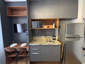 A compact kitchen with a microwave, sink, and refrigerator. There is a wooden shelf above the microwave and a dining table with chairs.