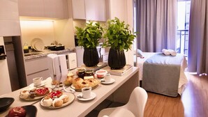 A modern hotel room with a dining area featuring a long table with various pastries and a coffee station, a small seating area with bar stools, a bed with a grey blanket, and a large window with curtains. - Prime Stay - Vila Buarque - São Paulo - BR (São Paulo)
