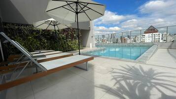 Piscina no terraço com espreguiçadeiras e guarda-sóis, vista para a cidade e fachada de um edifício moderno.