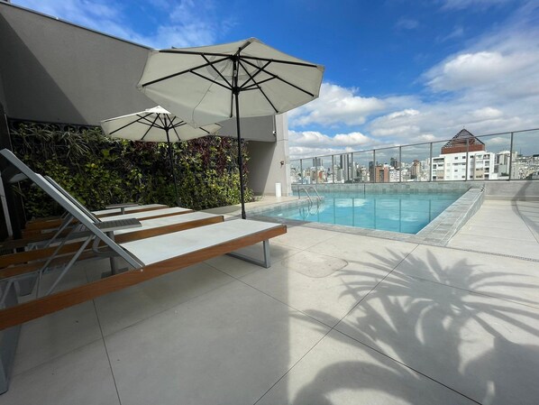 A rooftop pool with lounge chairs and umbrellas, a city view, and a modern building facade.
