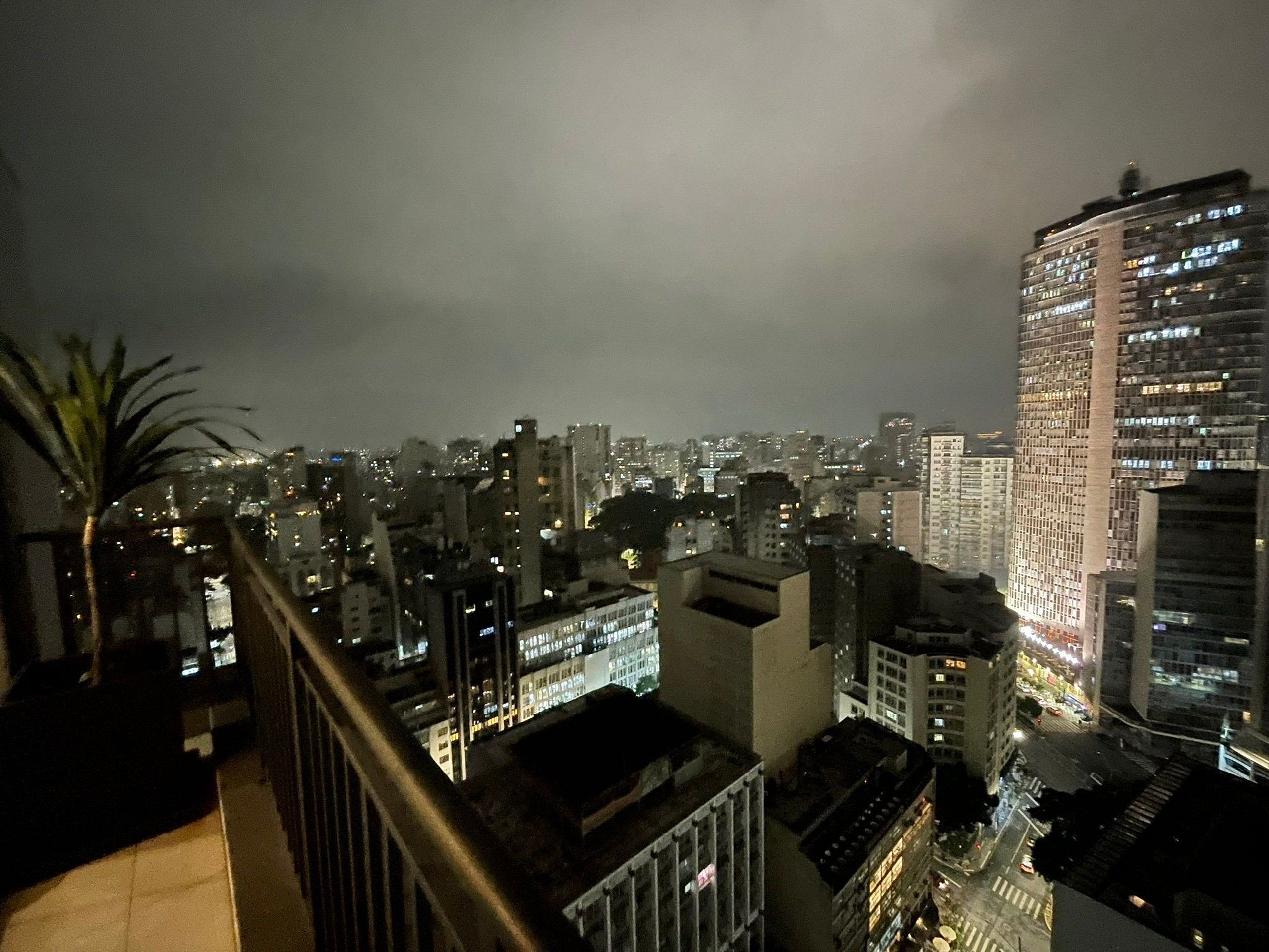 A nighttime cityscape with high-rise buildings, a palm tree, and a balcony railing.