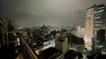 A nighttime cityscape with high-rise buildings, a palm tree, and a balcony railing.