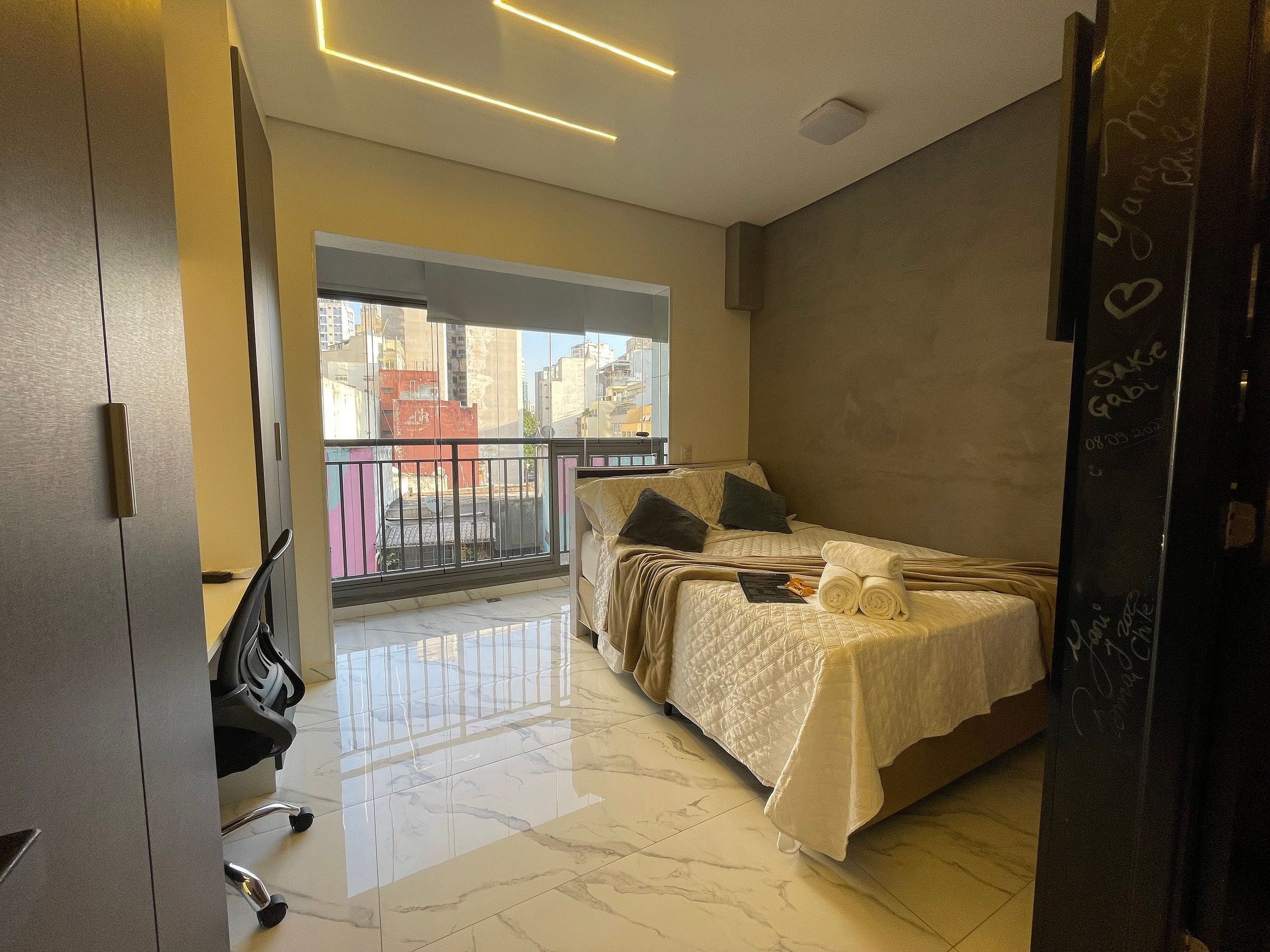 A modern bedroom with a bed, a desk, and a balcony with a city view.