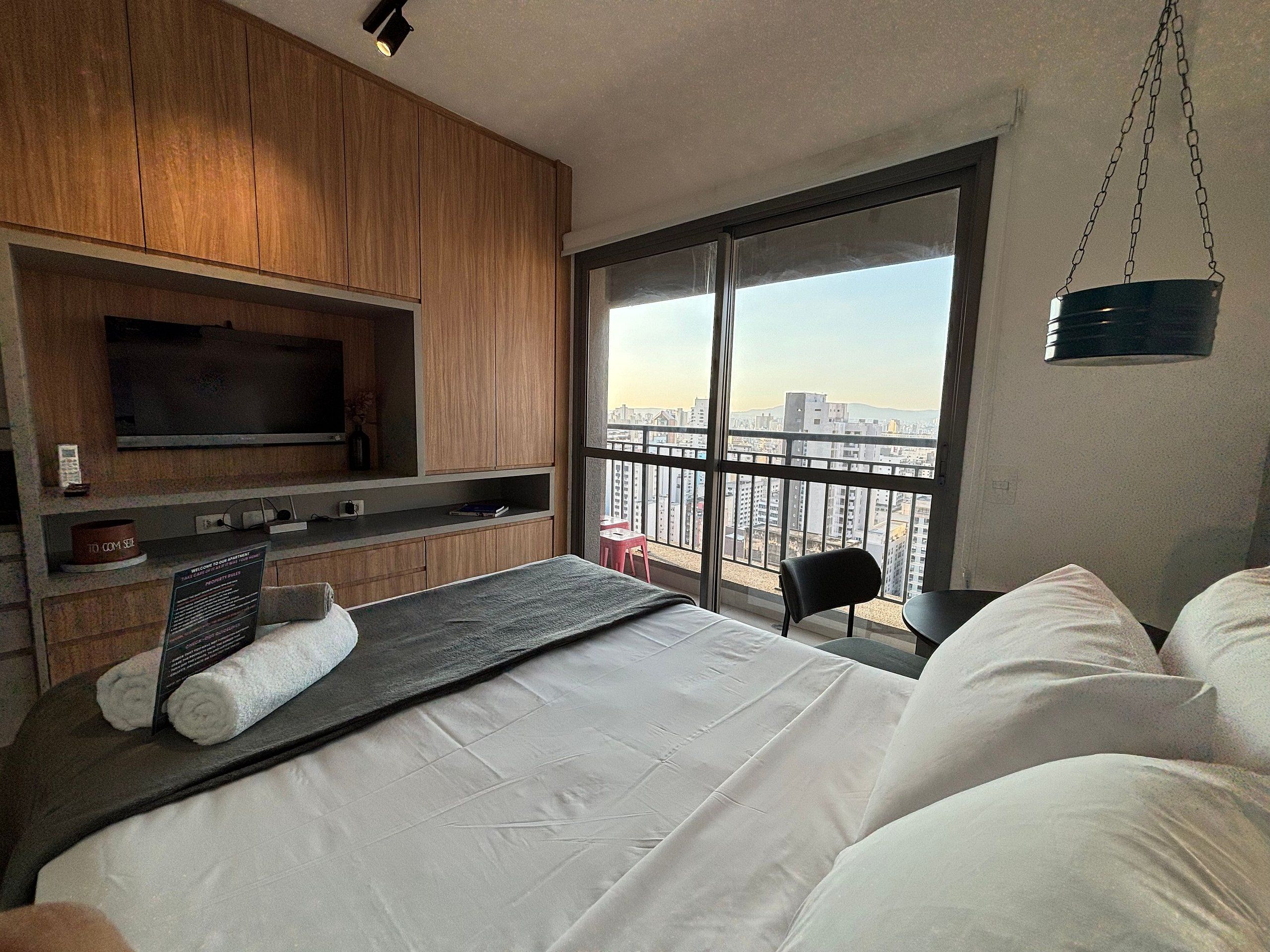 Une chambre d’hôtel moderne dotée d’un grand lit, d’une télévision à écran plat, d’un balcon offrant une vue sur la ville et d’un luminaire suspendu.
