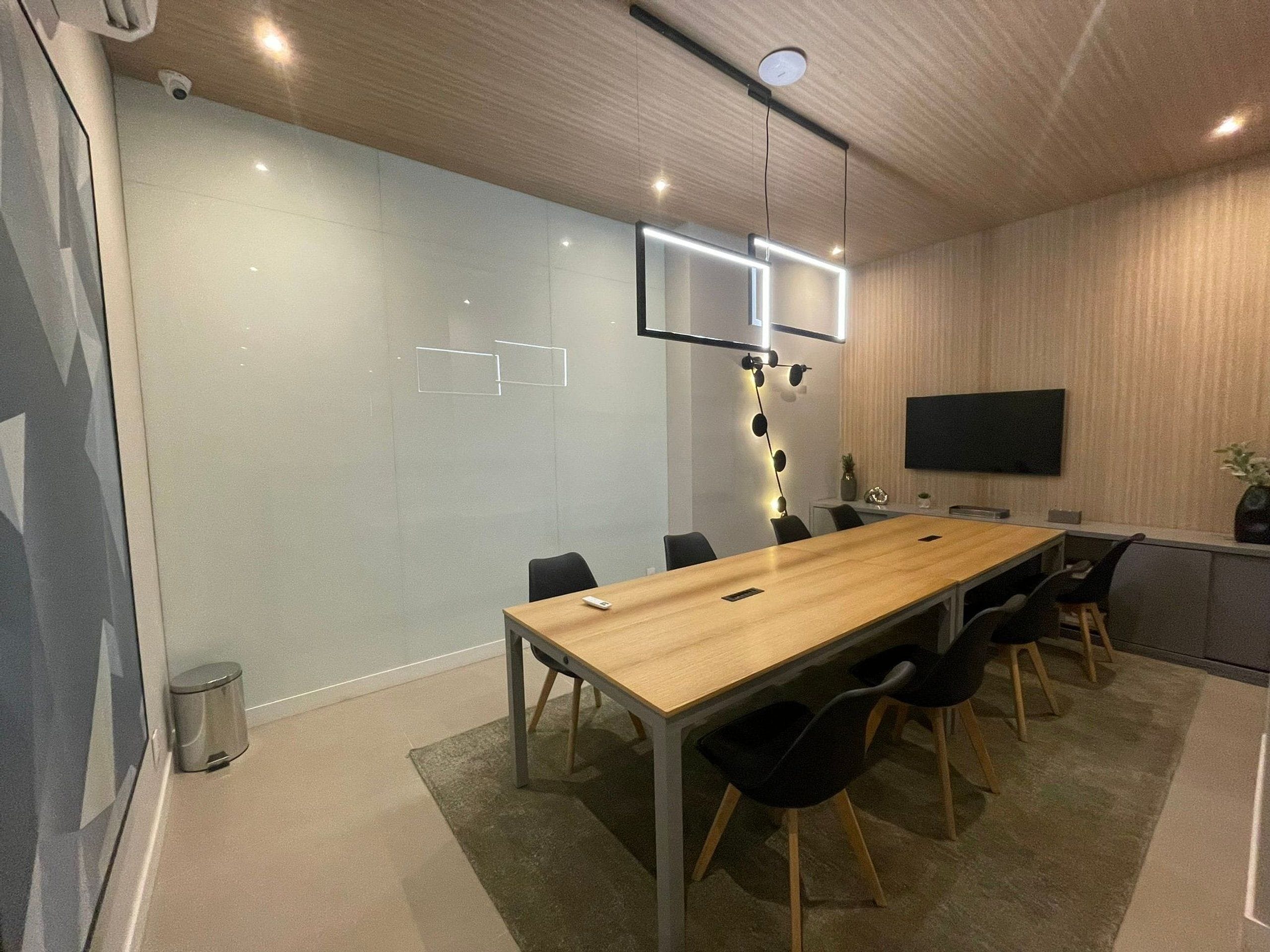 Une salle de conférence moderne, dotée d’une longue table en bois, de chaises noires, d’un téléviseur fixé au mur et d’un luminaire suspendu au plafond.