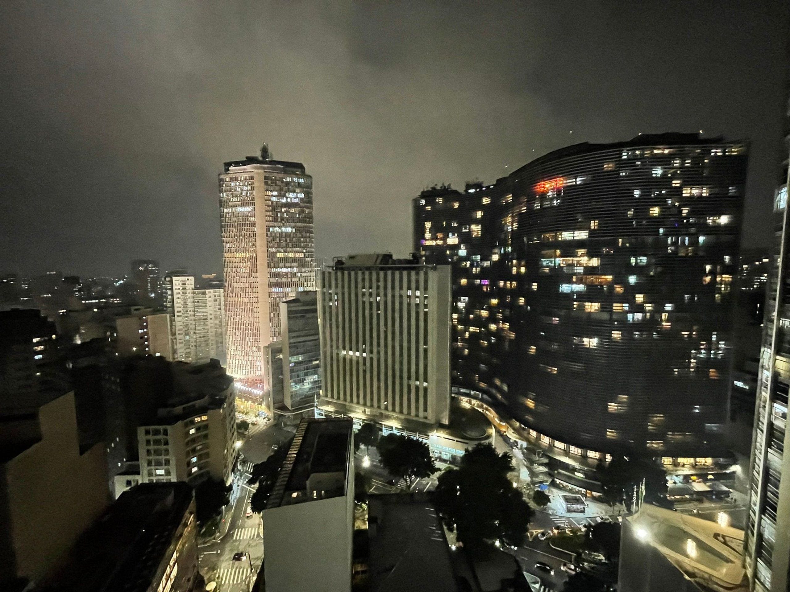 Une vue nocturne d’une ville, avec de hauts immeubles, dont l’un se distingue par sa façade incurvée et l’autre par son architecture verticale.