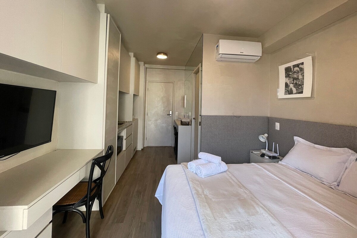 A hotel room with a bed, a desk with a chair, a television, a bedside table, and an air conditioning unit.