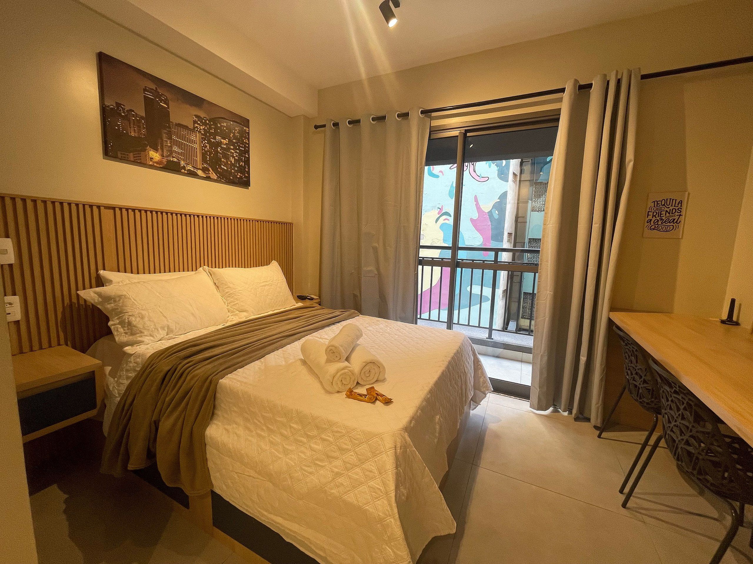 A hotel room with a bed, a desk, a balcony with curtains, and a cityscape painting on the wall.