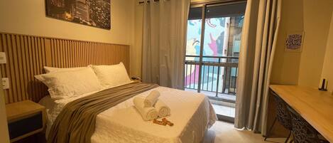 A hotel room with a bed, a desk, a balcony with curtains, and a cityscape painting on the wall.
