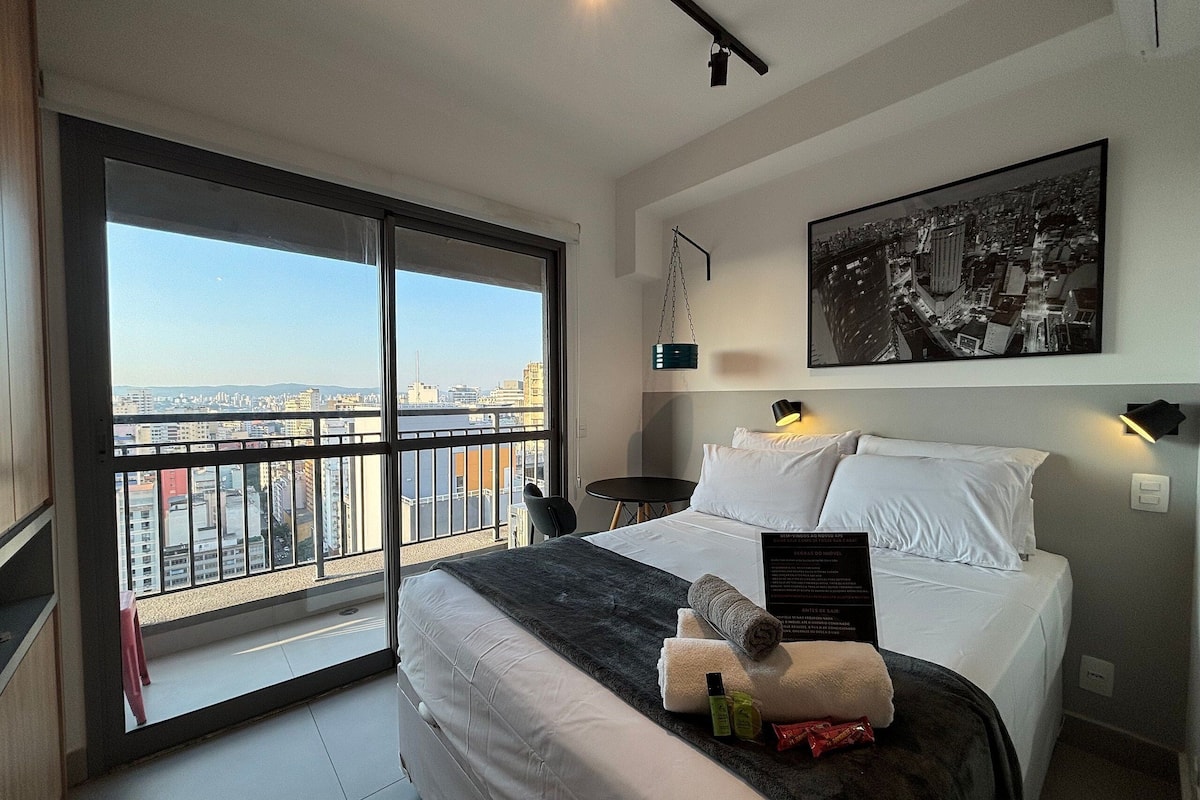 Hotelkamer met een groot bed, stadszicht door een schuifdeur, een ingelijst stadsbeeld en een klein tafeltje met een stoel.