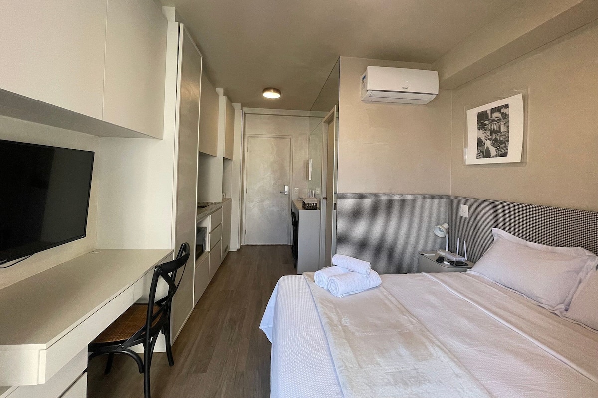 Een hotelkamer met een bed, een bureau met stoel, een televisie, een nachtkastje en een airconditioner.