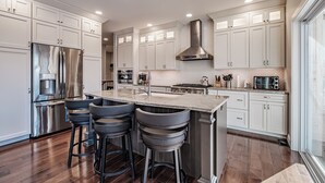 A modern kitchen with white cabinetry, stainless steel appliances, a central island with bar stools, and a large window providing natural light. - Beautiful home located on the coveted Paradise Road in Bethel Maine (Bethel)
