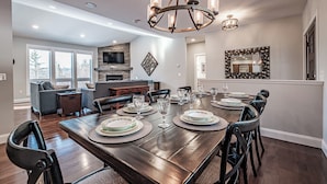 A spacious dining area with a long wooden table set for six, surrounded by dark chairs. A modern chandelier hangs above, and a stone fireplace is visible in the background. - Beautiful home located on the coveted Paradise Road in Bethel Maine (Bethel)