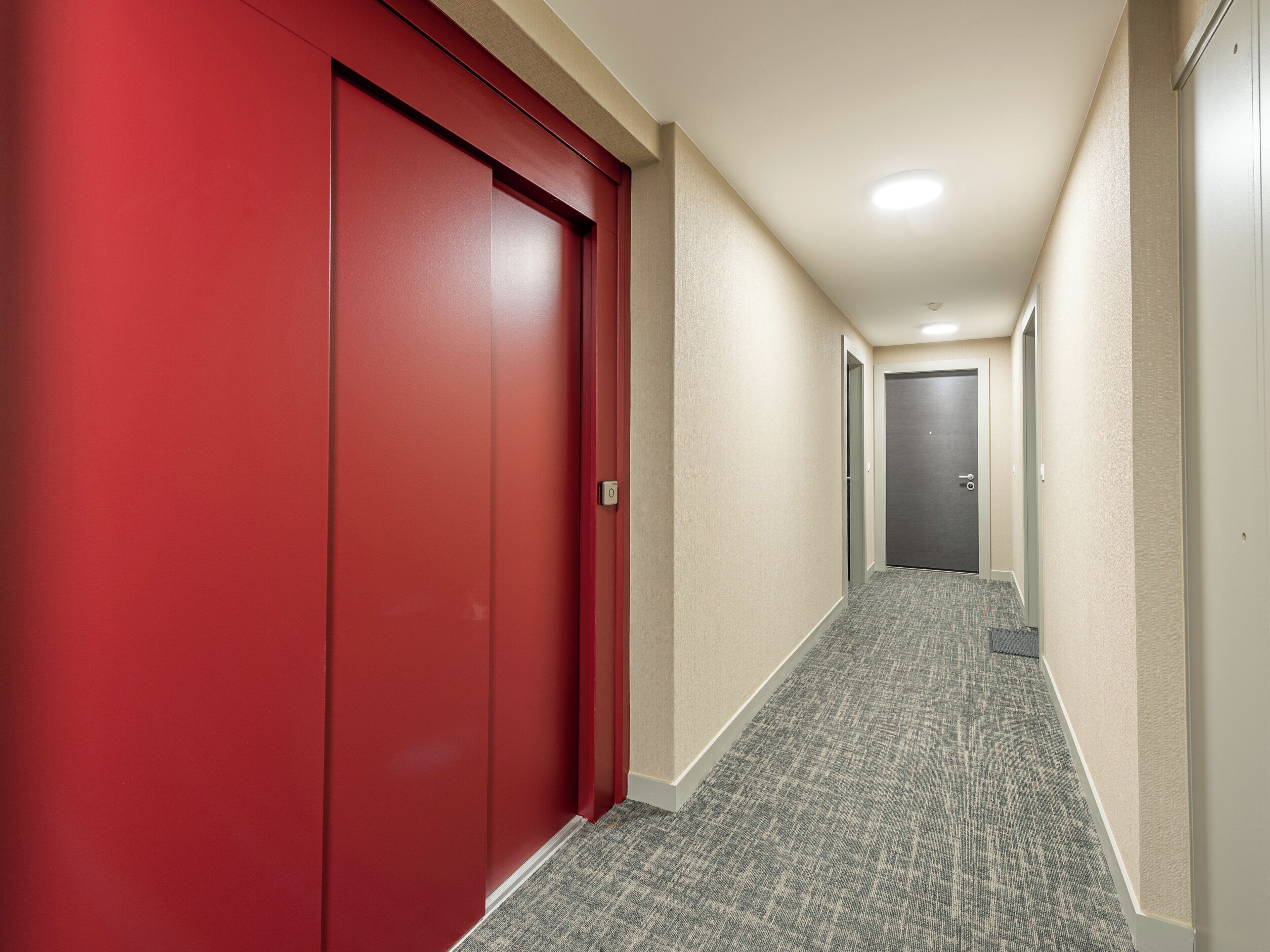 Un couloir avec une porte rouge sur la gauche et une porte grise au fond. Les murs sont de couleur beige et le sol est recouvert d’une moquette.