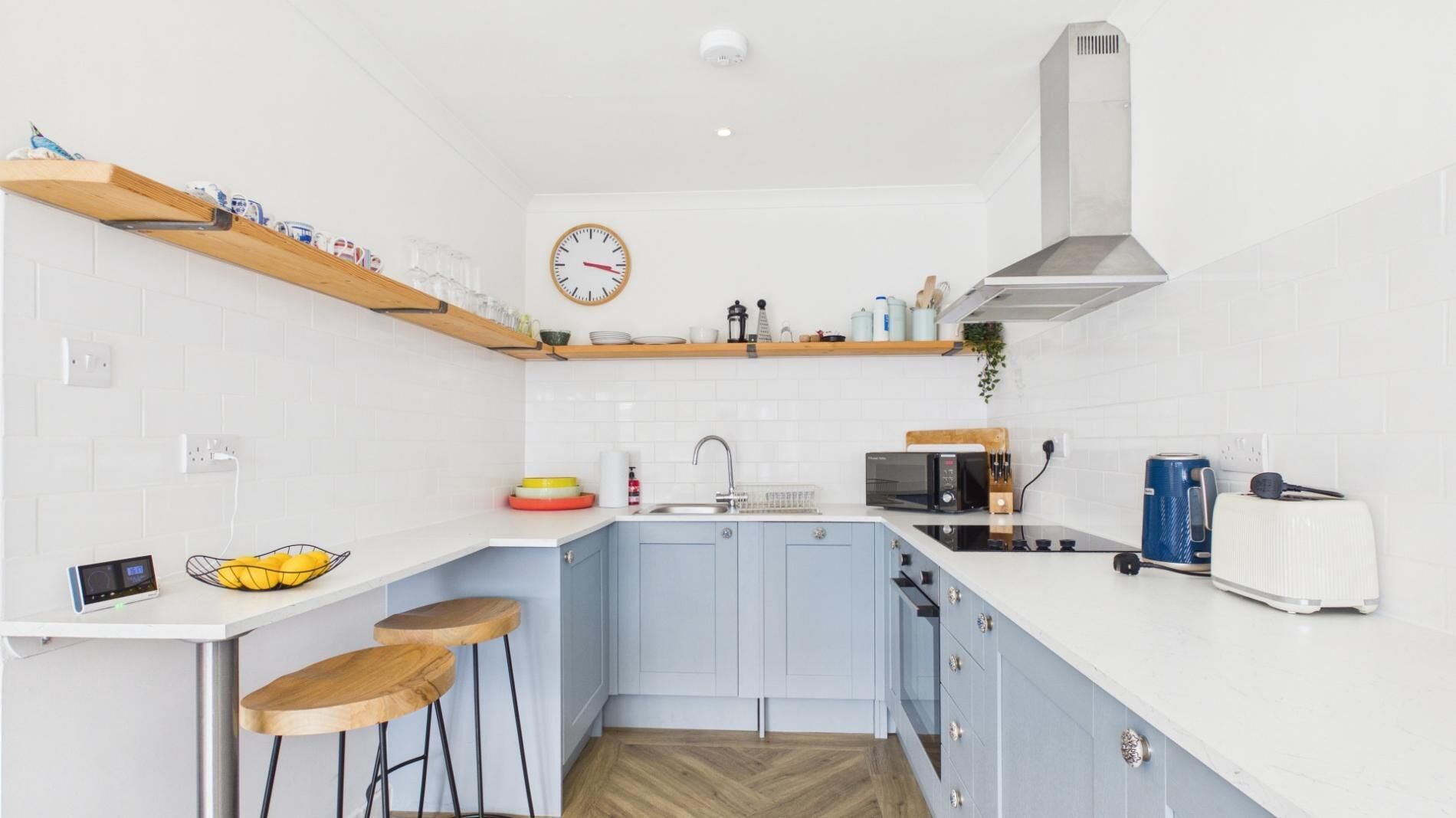 Een moderne keuken met witte en blauwe kastjes, een houten bar en een magnetron, toaster en een klok aan de muur.