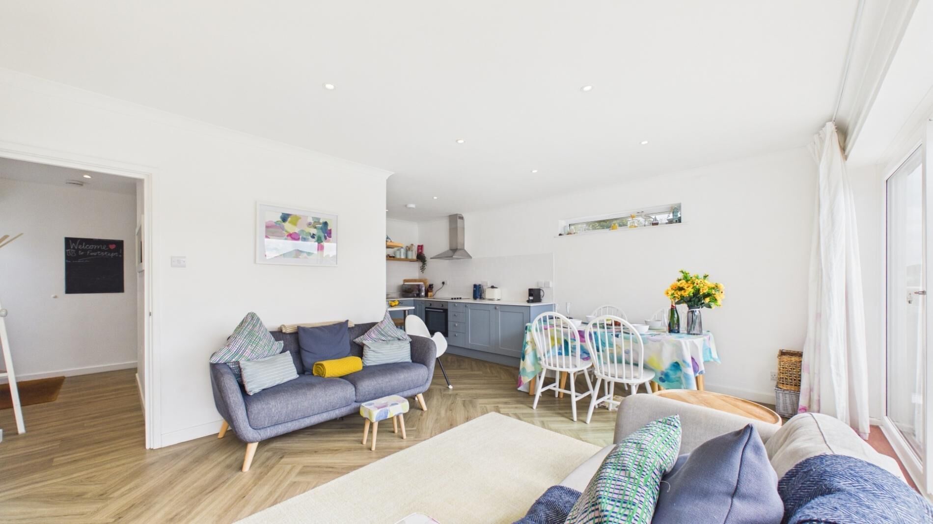 A modern living room with a sofa, a dining table with chairs, a kitchen area, and a large window.