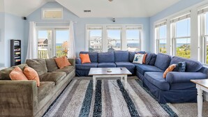 A living room with a sectional sofa, a coffee table, and a ceiling fan. There are large windows with a view of a house and the ocean. - Calypso: Semi-Soundfront w/ Pool | Wind Over Waves (Salvo)