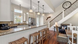 A spacious kitchen with a central island, bar stools, and a staircase leading to a dining area. - Vitamin Sea: Hot Tub | Rec Room | Pool Access (Corolla)