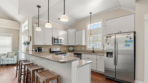 A modern kitchen with white cabinetry, a stainless steel refrigerator, a granite countertop, and pendant lighting. - Vitamin Sea: Hot Tub | Rec Room | Pool Access (Corolla)