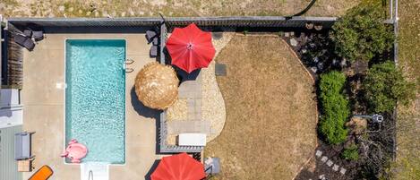 Vista aérea de un patio trasero con piscina, dos sombrillas rojas, un jacuzzi y una fogata.