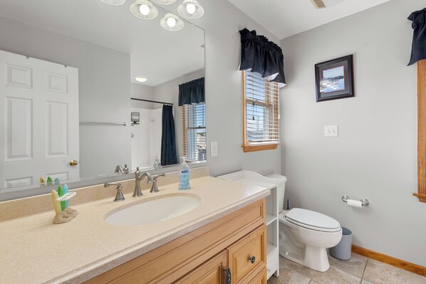 A bathroom with a sink, mirror, toilet, and window with blinds.