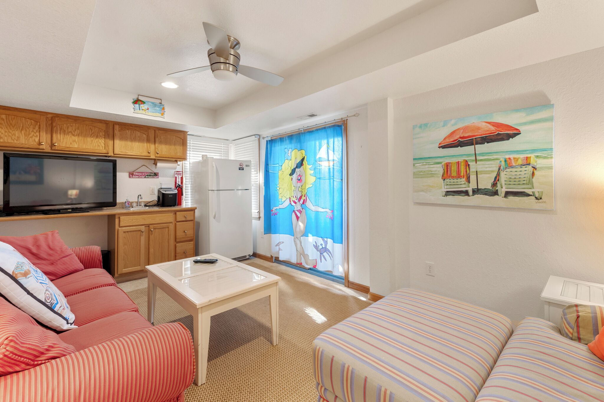 Un salon avec un canapé, un téléviseur, un ventilateur de plafond et un tableau représentant des chaises de plage.
