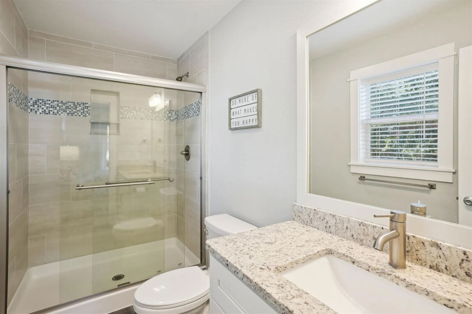 Une salle de bain dotée d’une douche à parois de verre, de toilettes, d’un lavabo avec un plan de travail en granit et d’un miroir placé au-dessus d’une fenêtre.